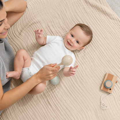 Happy baby playing with a wooden rattle on a cozy muslin cotton blanket with mom nearby.