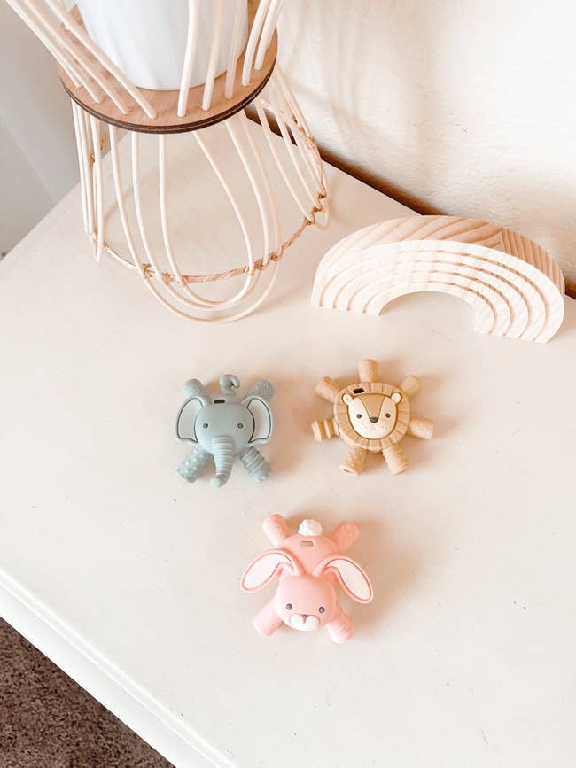 Pastel animal-shaped teethers including a pink bunny, grey elephant, and beige lion on a white surface with decor.