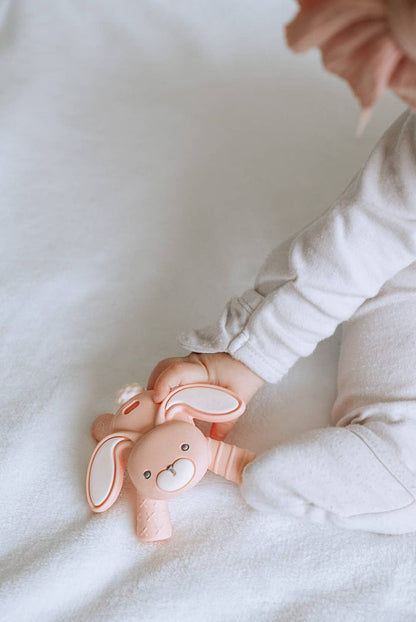 Baby holding Itzy Ritzy Ritzy Teether Bunny, offering soothing teething relief with eco-friendly design on soft white blanket.